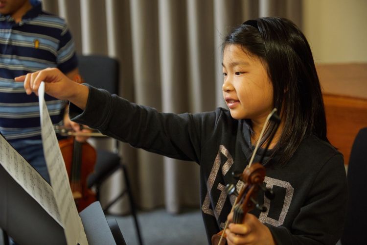 A girl with black hair and long sleeved black top sits holding a violin with her left hand, whilst turning a page of sheet music with her right hand